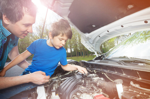 Portrait of little boy sitting on car hood and watching his father work on engine outdoors - Photo, Image