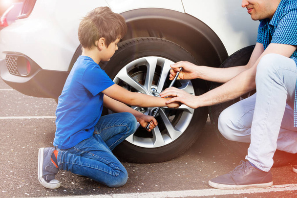Portrait of little boy and his father changing wheels on car with cross wheel wrench outside - Foto, imagen