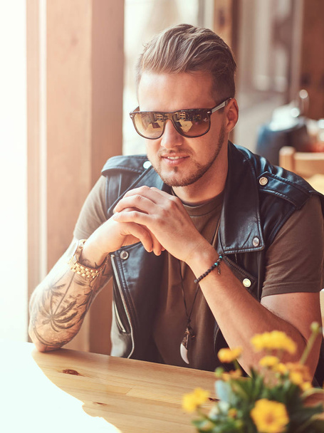 Hipster con un corte de pelo elegante y barba se sienta en una mesa en un café al lado de la carretera
. - Foto, Imagen
