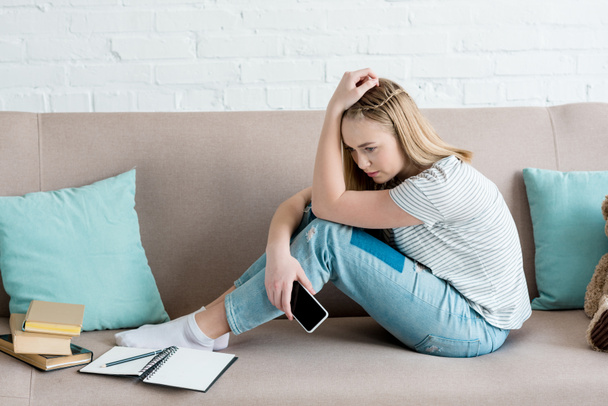 depressives Teenager-Mädchen sitzt mit Smartphone auf Couch, während sie Hausaufgaben macht - Foto, Bild