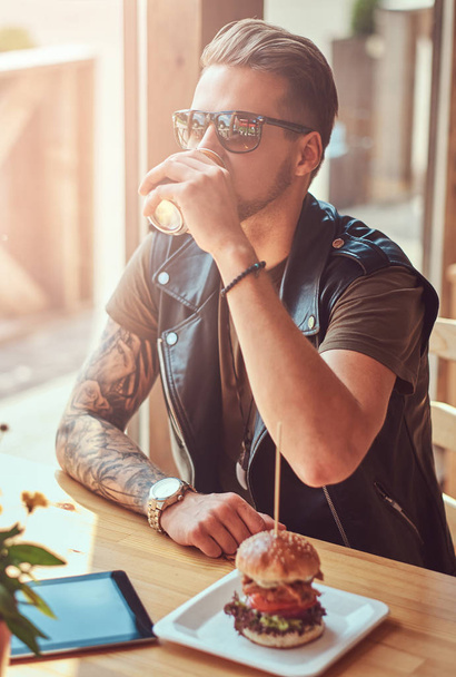 Hipster con un corte de pelo elegante y barba se sienta en una mesa, se detuvo a almorzar en un café al lado de la carretera
. - Foto, imagen