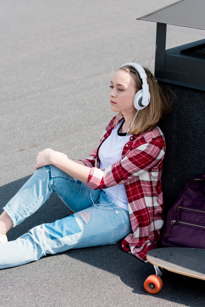 όμορφη έφηβος κορίτσι με skateboard κάθεται στον τελευταίο όροφο και να ακούει μουσική με ακουστικά - Φωτογραφία, εικόνα