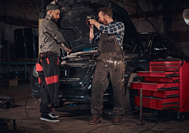 Dos barbudos mecánico de automóviles en un uniforme, reparar un coche roto en el garaje
.  - Foto, imagen