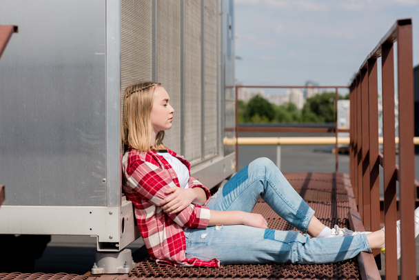 Seitenansicht des nachdenklichen Teenie-Mädchens im rot karierten Hemd auf dem Dach sitzend - Foto, Bild
