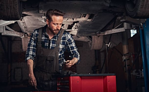 Auto mechanic in a uniform, working on a workbench while standing under lifting car in a repair garage.  - Photo, image