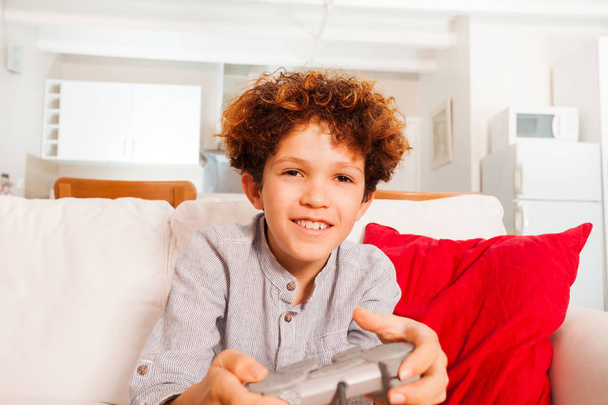 Close-up portrait of curly preteen boy with joystick playing video games in the living room - Фото, изображение