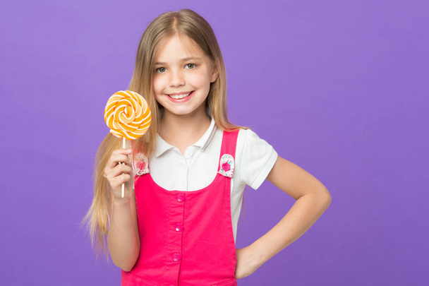Girl eating big candy on stick or lollipop. Sweet childhood concept. Kid with long hair likes sweets and treats. Girl on smiling face holds giant colorful lollipop in hand, violet background - Photo, image