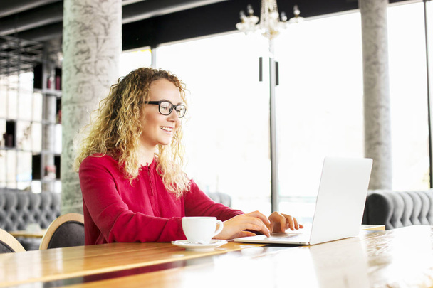 Happy beautiful curly hair blonde young female at coffee shop using laptop, smiling. Young attractive woman freelancer wearing glasses surfing web on hipster notebook computer. Background, copy space. - Foto, Bild