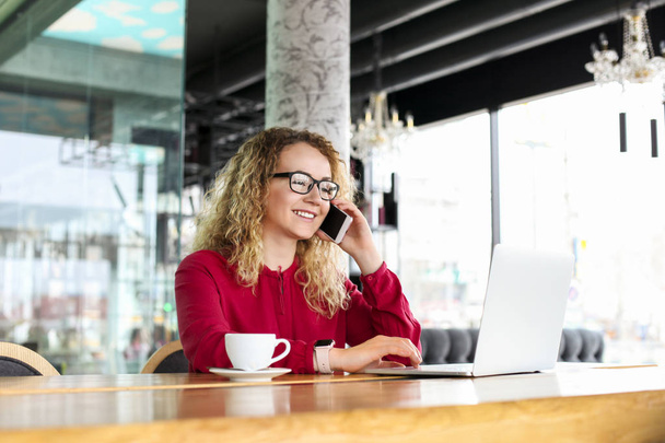 Attractive young woman sitting at coffee shop with her fashionable laptop, drinking cappuccino, talking on the phone, receiving good news smiling. Beautiful blonde female with curly hair. Background. - Photo, image
