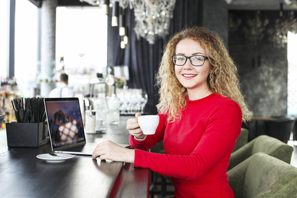 Beautiful blonde young woman drinking cappuccino coffee in a hipster bar and smiling. Curly haired female blogger, smiling, surfing, browsing, blogging on laptop computer. Windows background, portrait - Zdjęcie, obraz
