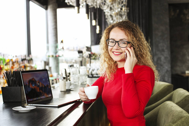 Ελκυστική νεαρή γυναίκα κάθεται στο καφενείο με το laptop της μόδας, πίνοντας καπουτσίνο, μιλάτε στο τηλέφωνο, λαμβάνουν καλές ειδήσεις χαμογελώντας. Όμορφη ξανθιά γυναίκα με σγουρά μαλλιά. Φόντο. - Φωτογραφία, εικόνα