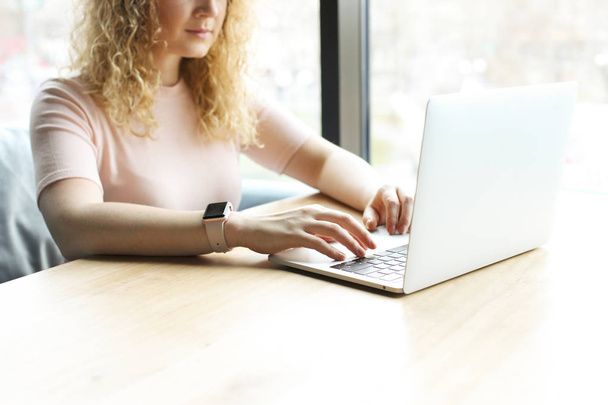 Cropped shot of attractive blonde young woman, sitting in coffee shop typing on blank screen laptop. Curly blonde female freelancer working, browsing on her notebook computer. Background, close up. - Fotó, kép