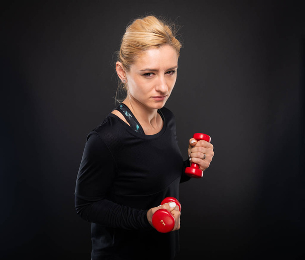 Pretty gym female training arms with dumbbells looking mad on black background - Zdjęcie, obraz