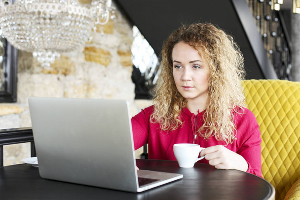 Όμορφη ξανθιά νεαρή γυναίκα πίνοντας καφέ καπουτσίνο σε ένα hipster bar και χαμογελαστός. Σγουρά μαλλιά γυναίκα blogger, χαμογελώντας, το σερφ, περιήγηση, blogging στον φορητό υπολογιστή. Φόντο των Windows, πορτρέτο - Φωτογραφία, εικόνα