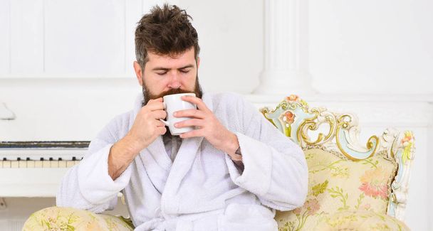 Man sleepy in bathrobe, drinks coffee, enjoying aroma in luxury hotel in morning, white background. Elite leisure concept. Man with beard and mustache enjoys morning while sitting on luxury armchair. - Photo, image