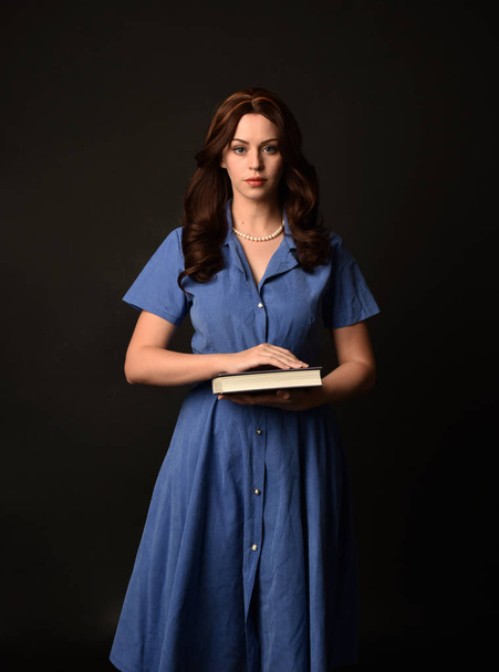 3/4 portrait of brunette lady wearing blue dress, holding a book. posed on black studio background. - Photo, image