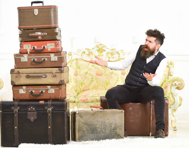 Macho elegant on surprised face sits shocked near pile of vintage suitcase. Luggage and travelling concept. Man, butler with beard and mustache delivers luggage, luxury white interior background - Photo, image