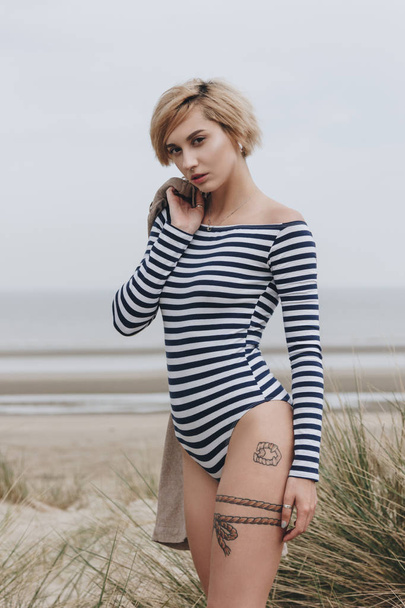 beautiful young woman in striped bodysuit on sandy shore on cloudy day - Фото, зображення