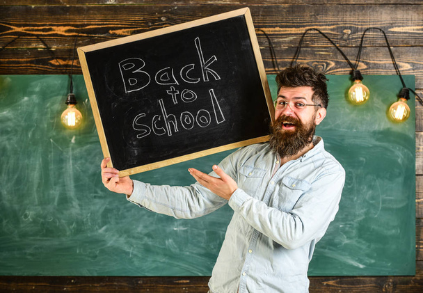 Teacher in eyeglasses holds blackboard with title back to school. Man with beard and mustache on happy face welcomes students, chalkboard on background. High school concept - Фото, зображення