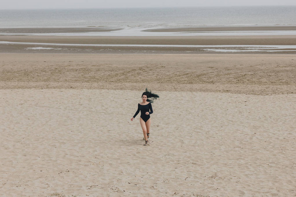 attractive young woman in black bodysuit running on sandy seashore - Фото, изображение