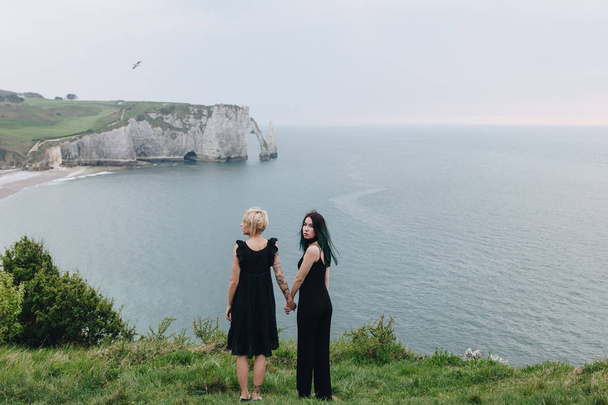 Rückansicht junger Frauen, die sich an bewölkten Tagen an einer Klippe vor dem Meer die Hände reichen - Foto, Bild