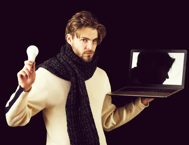 handsome bearded man or guy with beard on face in white sweater and knitted on black background holds laptop and lamp or light bulb - Fotó, kép