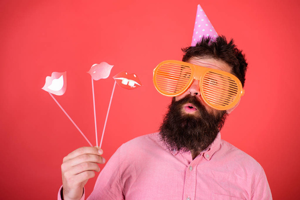 Emotional diversity concept. Hipster in giant sunglasses celebrating. Man with beard on cheerful face holds smiling lips on sticks, red background. Guy in party hat celebrate, posing with photo props - Foto, imagen