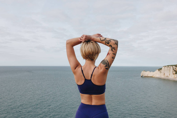 rear view of tattooed athletic girl in stylish sportswear posing near the sea, Etretat, France - Photo, Image