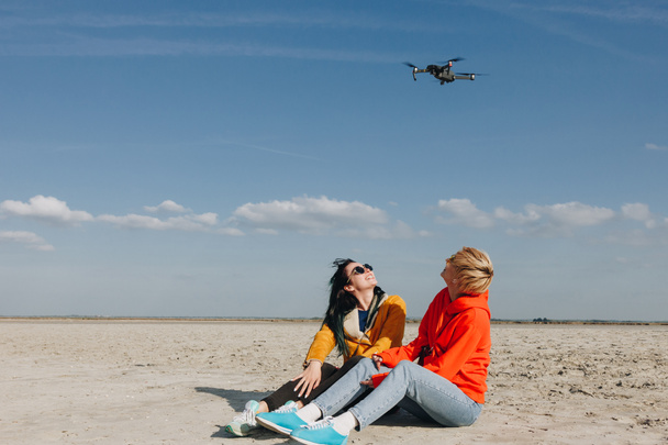 stijlvolle meisjes zittend op strand en kijken naar drone, mount Saint michaels, Normandië, Frankrijk - Foto, afbeelding