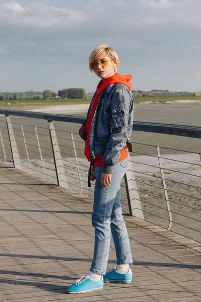 full length view of beautiful stylish girl in denim clothes and sunglasses looking at camera at sunny day, mont saint michel, france - Photo, Image