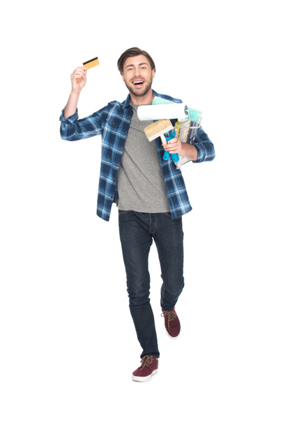 glücklicher Mann mit Kreditkarte mit Farbrollen und Pinsel isoliert auf weißem Hintergrund  - Foto, Bild