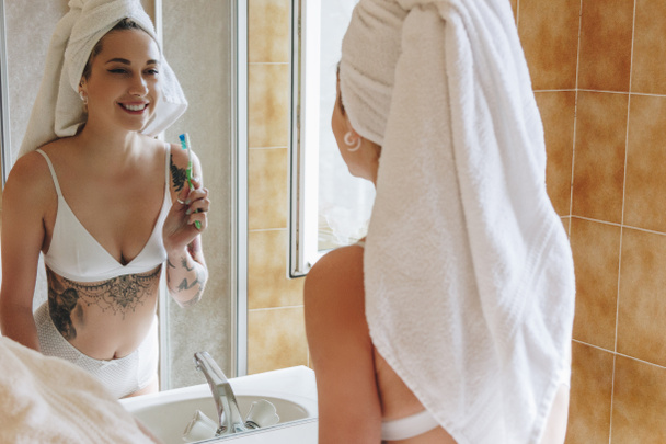 beautiful smiling young woman in underwear holding toothbrush and looking at mirror in bathroom - Photo, Image