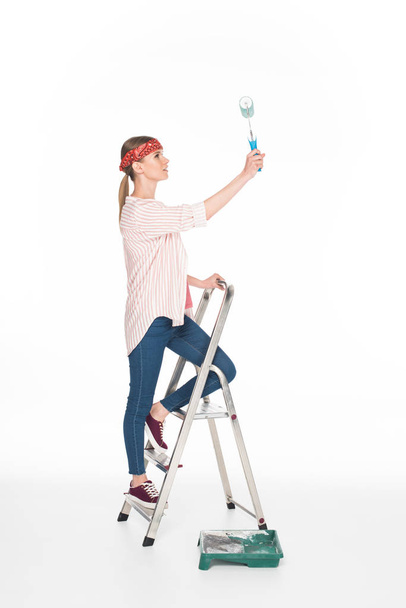 vue latérale de la femme dans le bandeau debout sur l'échelle et la peinture par rouleau de peinture isolé sur fond blanc
  - Photo, image