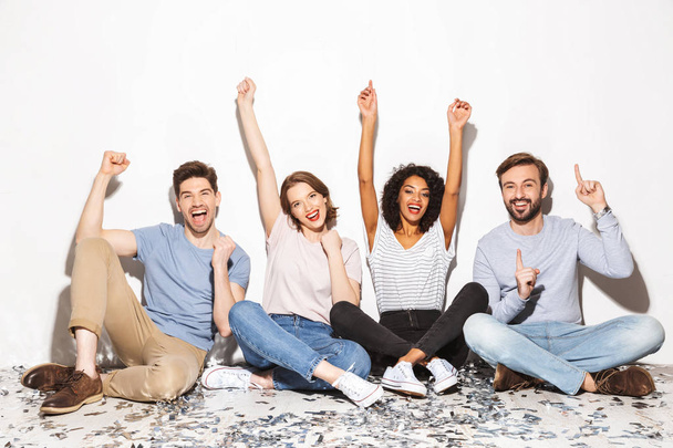 Group of happy multiracial people sitting on a floor with confetti and celebrating isolated over white background - Фото, изображение