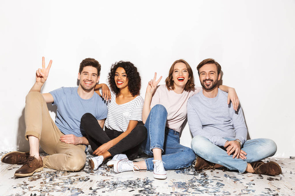 Group of happy multiracial people sitting on a floor with confetti isolated over white background - Φωτογραφία, εικόνα
