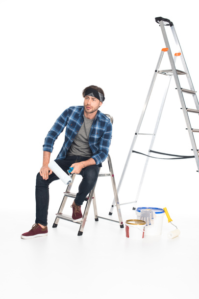 man in hoofdband zittend op de ladder met verfroller geïsoleerd op witte achtergrond  - Foto, afbeelding