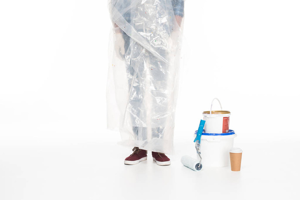 oříznutý obraz člověka v polyethylenu kryt stojící poblíž plechovek barvy, malování válečkem a šálek izolované na bílém pozadí  - Fotografie, Obrázek