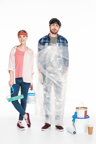 smiling woman with paint tin and paint roller standing with boyfriend in polyethylene cover near painting tools and coffee cup isolated on white background  - Photo, Image