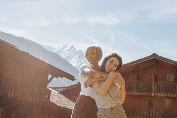 beautiful happy girls hugging while standing between wooden houses in mountain village, mont blanc, alps - Photo, Image
