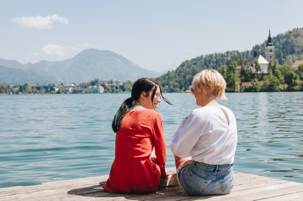 Achteraanzicht van stijlvolle meisjes zitten op houten pier in de buurt van rustige bergmeer, bled, Slovenië - Foto, afbeelding