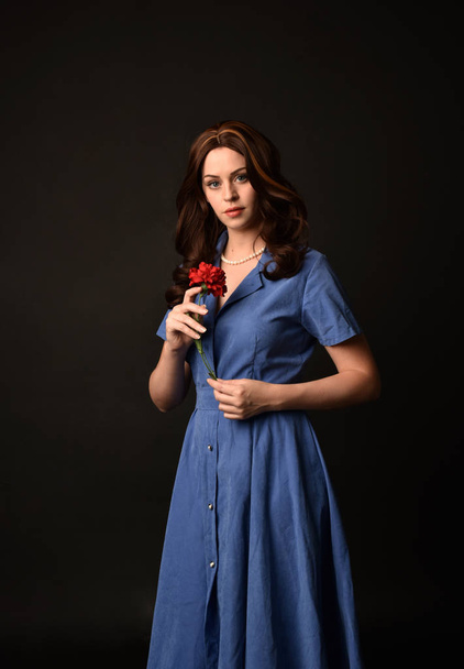 3/4 portret van brunette dame draagt blauwe jurk, houden van een bloem. gesteld op zwarte studio achtergrond. - Foto, afbeelding