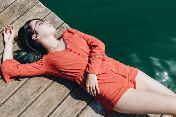 vista dall'alto di bella ragazza elegante in occhiali da sole sdraiato sul molo di legno vicino al lago
 - Foto, immagini
