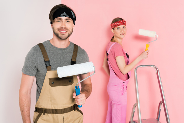 smiling man with paint roller and his girlfriend standing behind and painting wall  - Foto, Imagen