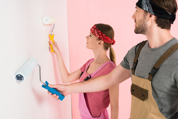 ペイント ローラーの間に壁を塗る作業のオーバー オールで若いカップルの側面図  - 写真・画像