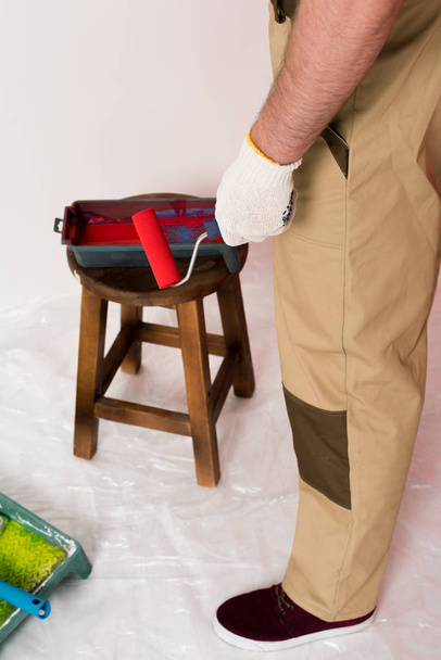 cropped shot of man in working overall holding paint roller and standing near roller tray on chair  - Photo, Image