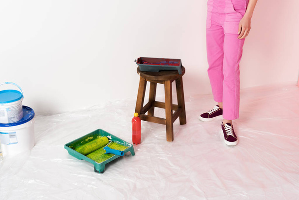 作業椅子、ローラー トレイ、ペンキ缶、ボトル近く全体に立っている女性のショットをトリミング  - 写真・画像