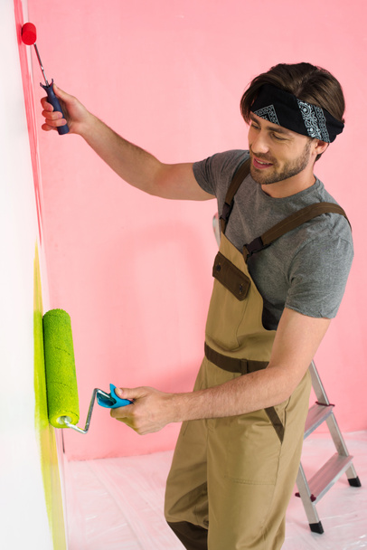 smiling man in working overall painting wall by two paint rollers  - Photo, Image
