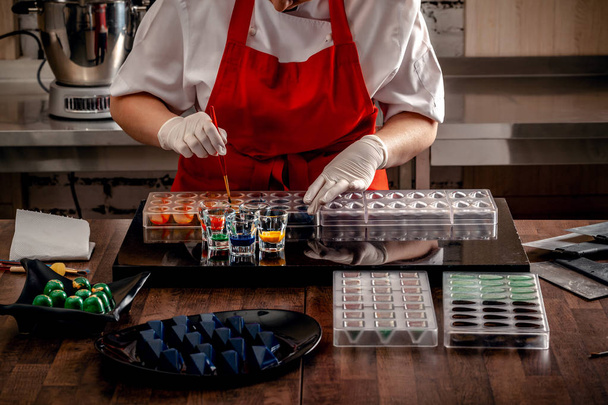 Kobieta cukiernik w czerwone rękawice jednolite i biały robi cukierki w specjalnych kształtach z mleczne czekolady z jadalne farby. - Zdjęcie, obraz