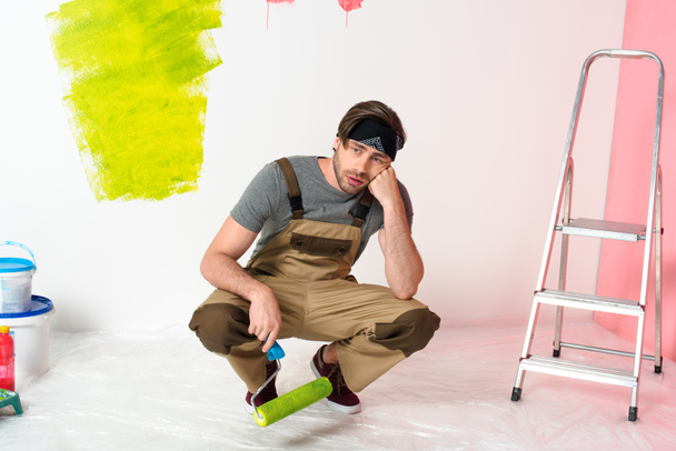 tired young man sitting with paint roller near ladder in front of painted wall  - Fotoğraf, Görsel