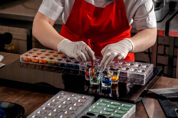 Femme un confiseur en uniforme rouge et gants blancs faisant des bonbons dans des formes spéciales de chocolat laitier avec de la peinture comestible
. - Photo, image